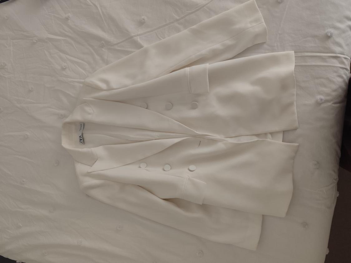 White button-up jacket on a bed with a textured white bedspread. Visible label with text "ZARA".