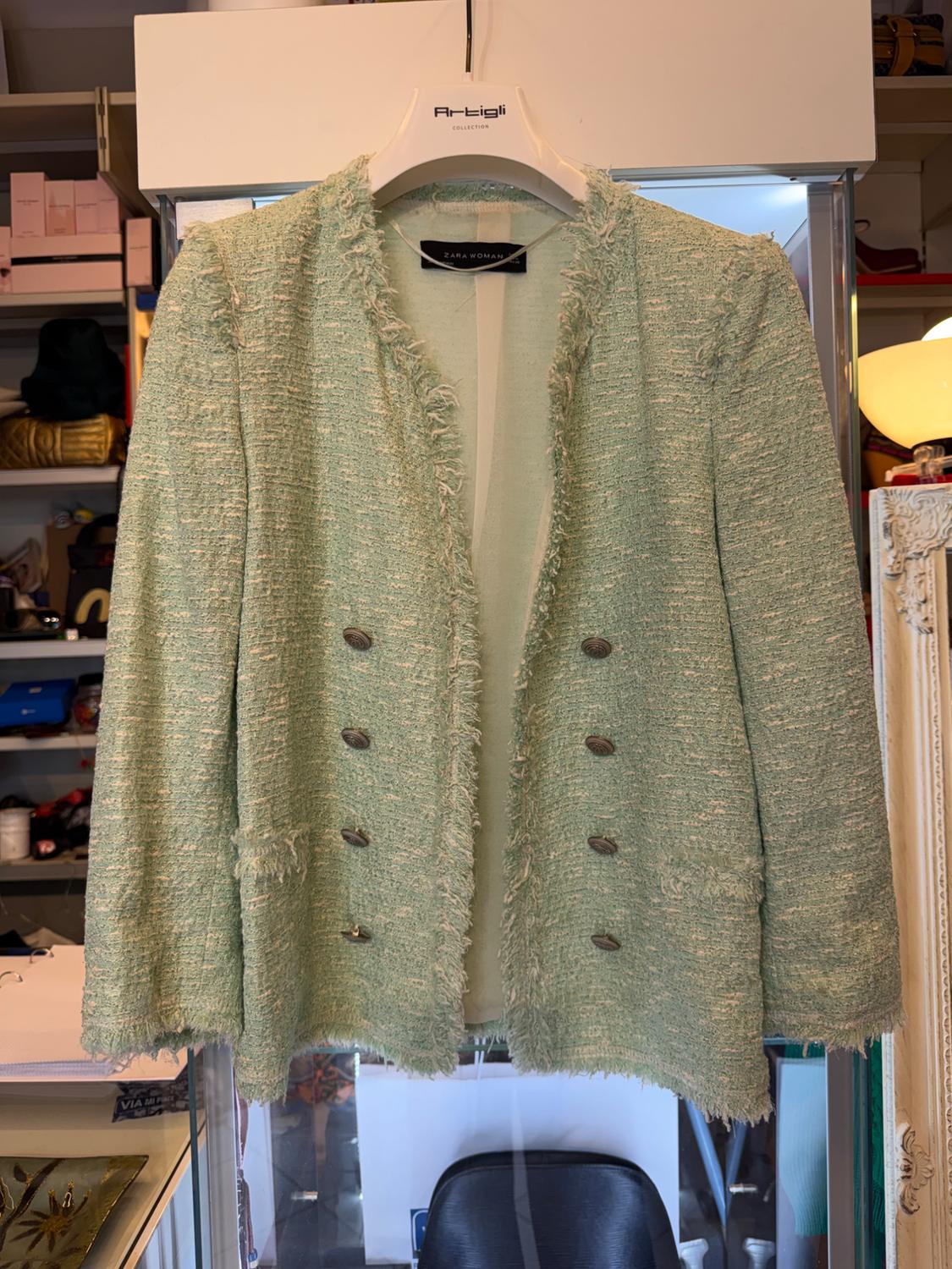 Light green textured jacket with buttons and frayed edges hanging in a room. Texts: Artigli COLLECTION, ZARA WOMAN.