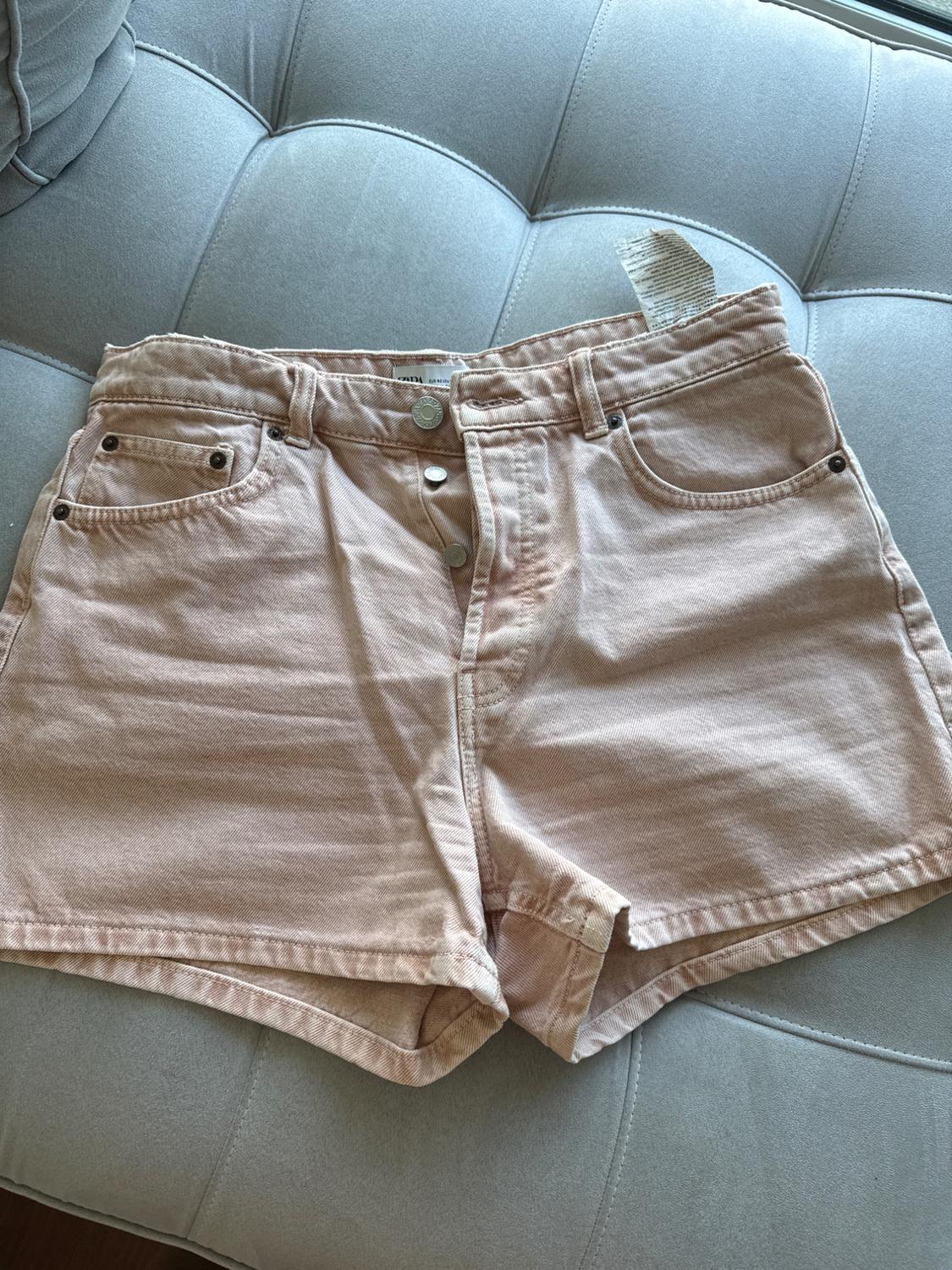 Light pink denim shorts with metallic buttons, on a light grey sofa. Label visible at the top.