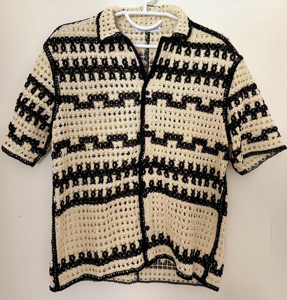 Black and white crochet shirt with a geometric pattern, hanging on a white hanger against a light background.