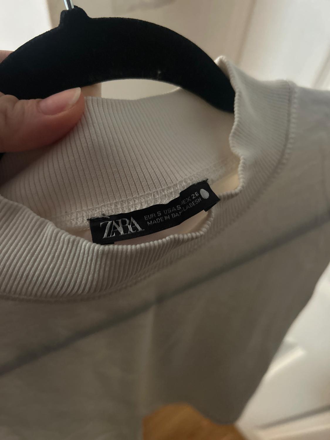 Hand holding a hanger with a white t-shirt. The label says "ZARA, EUR S USA S MEX 26, MADE IN BANGLADESH".