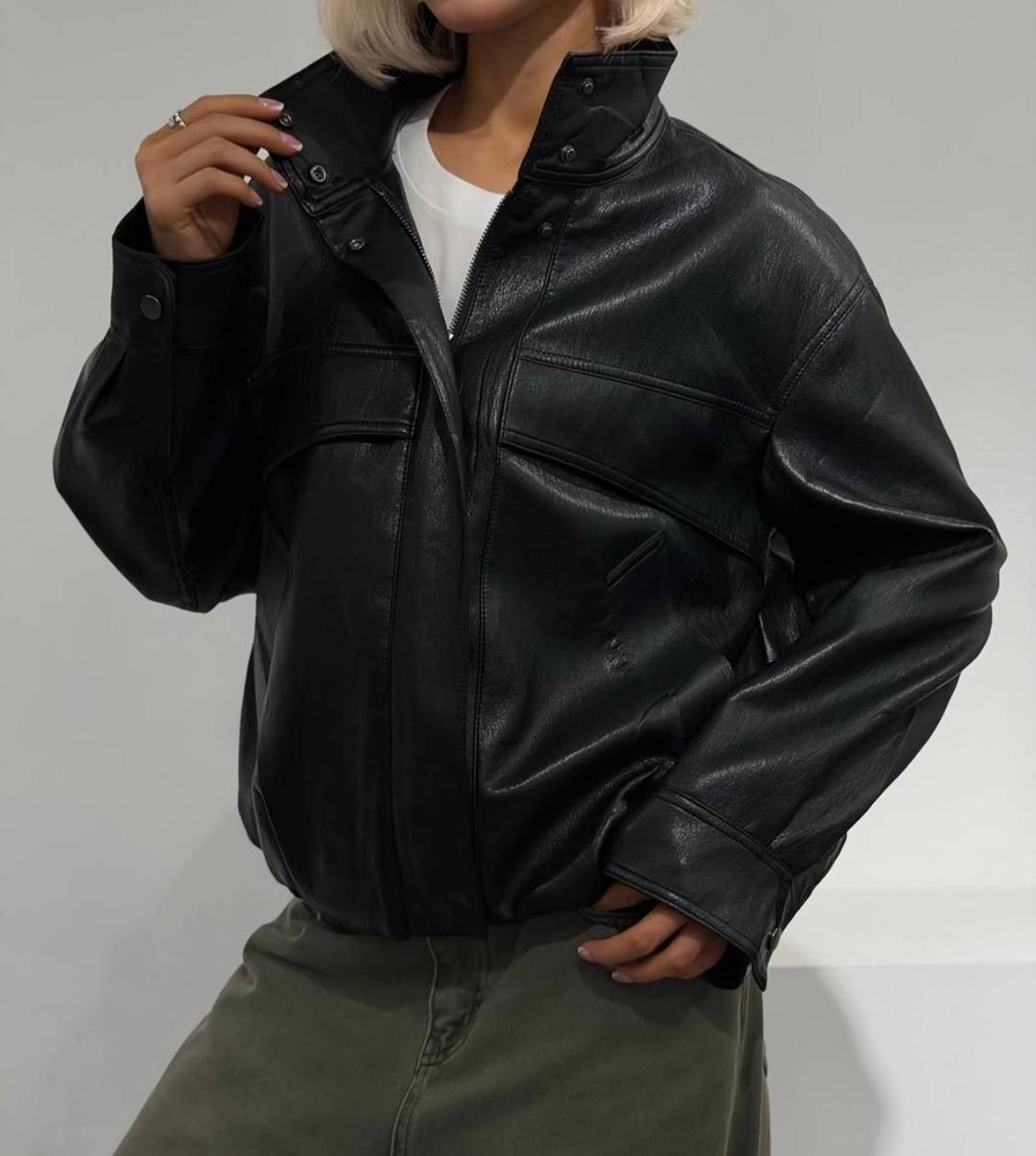 Blonde model wearing a black high-necked jacket, white t-shirt and olive green skirt against a light background. Casual style.