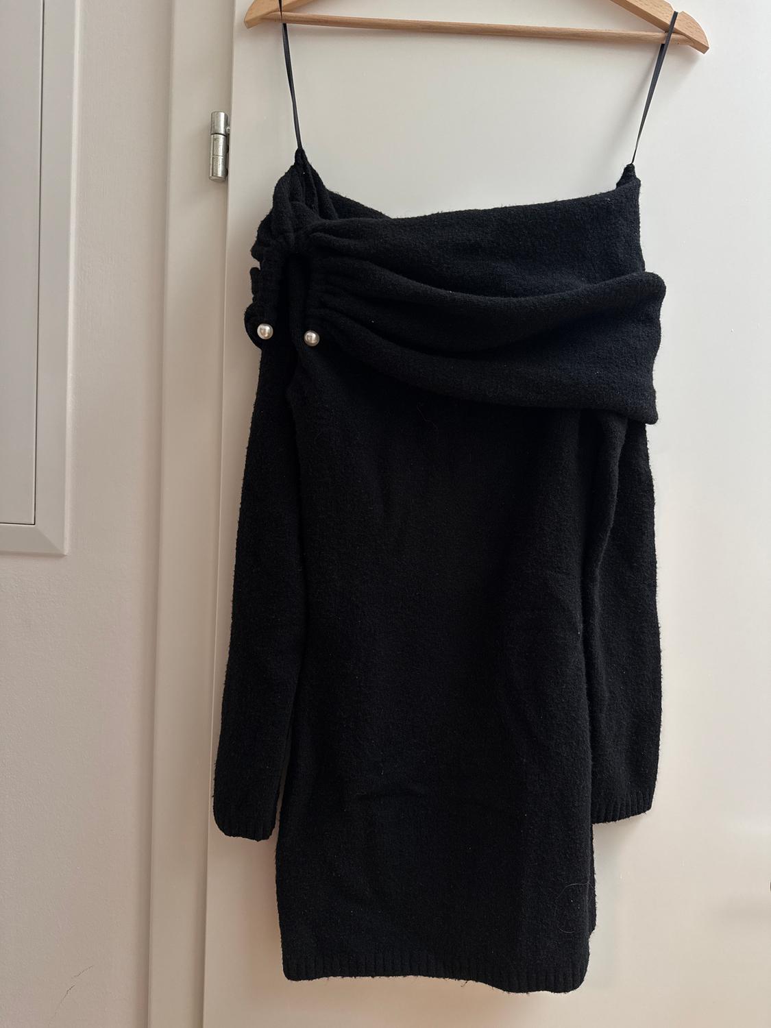 Black knitted dress with a draped neckline and pearl details on the shoulders, hanging on a wooden hanger.