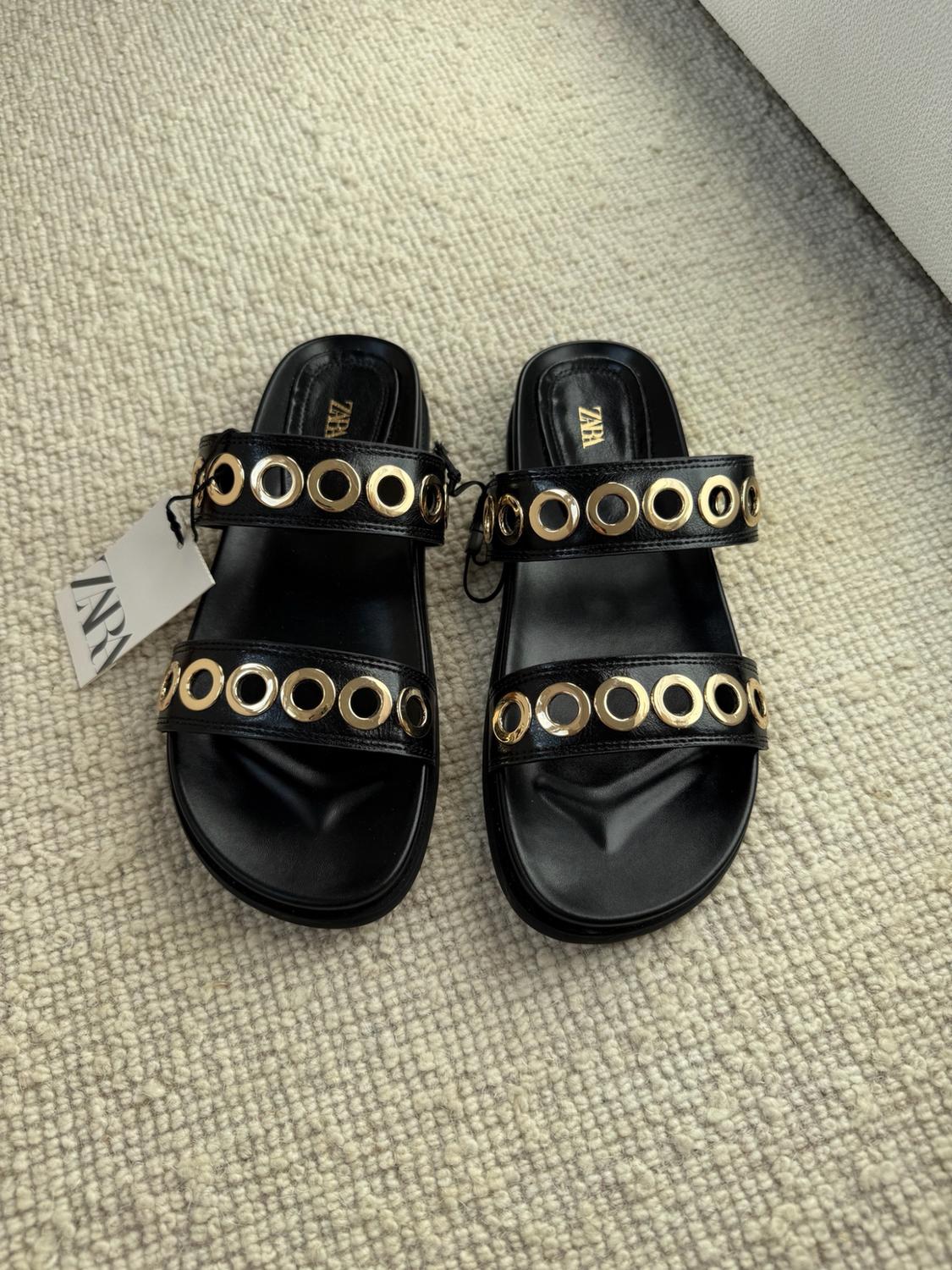 Black flat sandals with two straps and gold-toned rings. They feature a label with the text ZARA, set against a light textured rug.