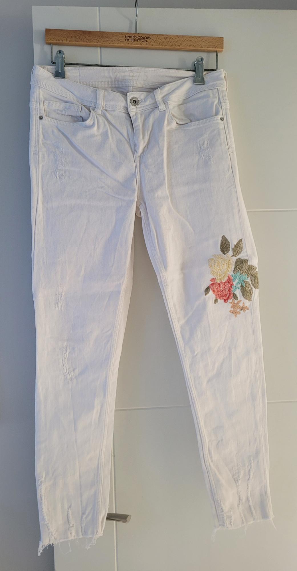 White trousers with floral embroidery on the right leg, hanging on a wooden hanger from United Colors of Benetton.