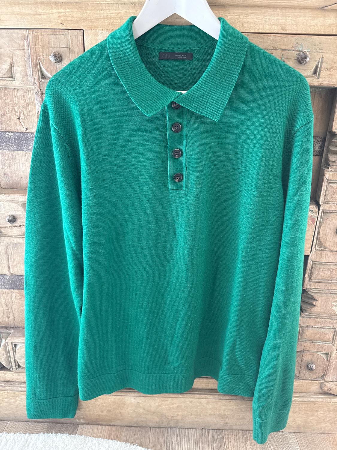 Green long-sleeved polo neck jumper with buttons, hanging on a white hanger in front of a wooden cabinet.