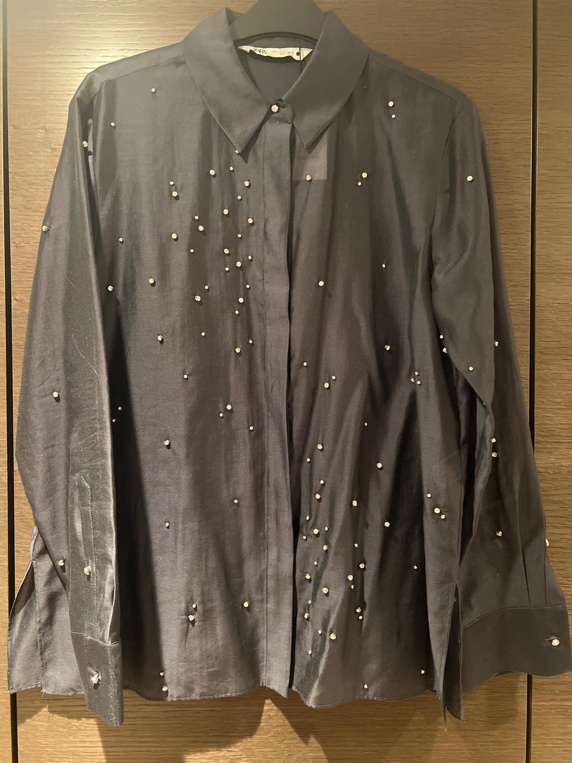 Black shirt with a classic collar and small decorative pearls, hanging on a hanger against a light wooden background.