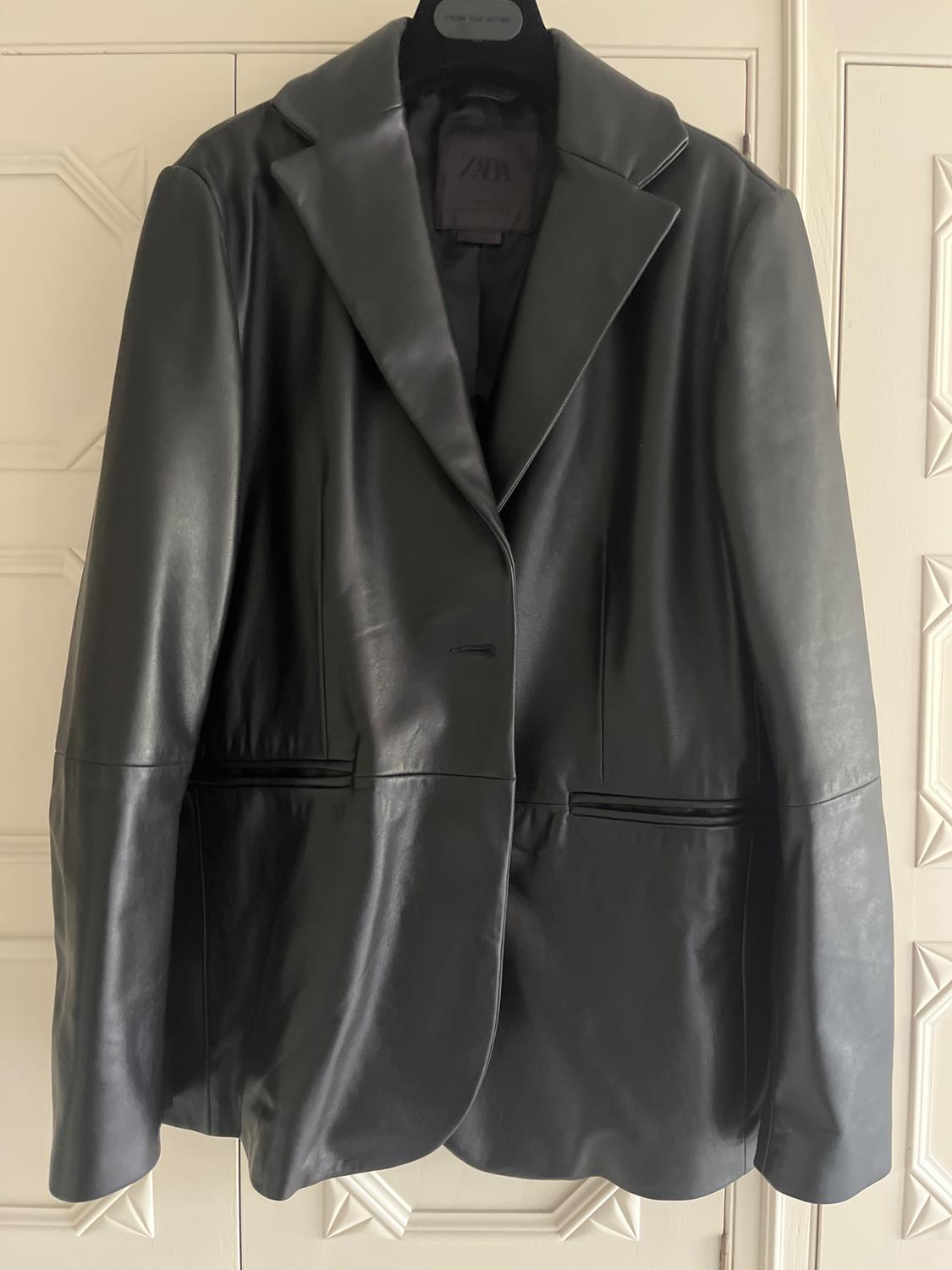 Black leather jacket hanging on a hanger, with wide lapels and front pockets, against a background of white doors.