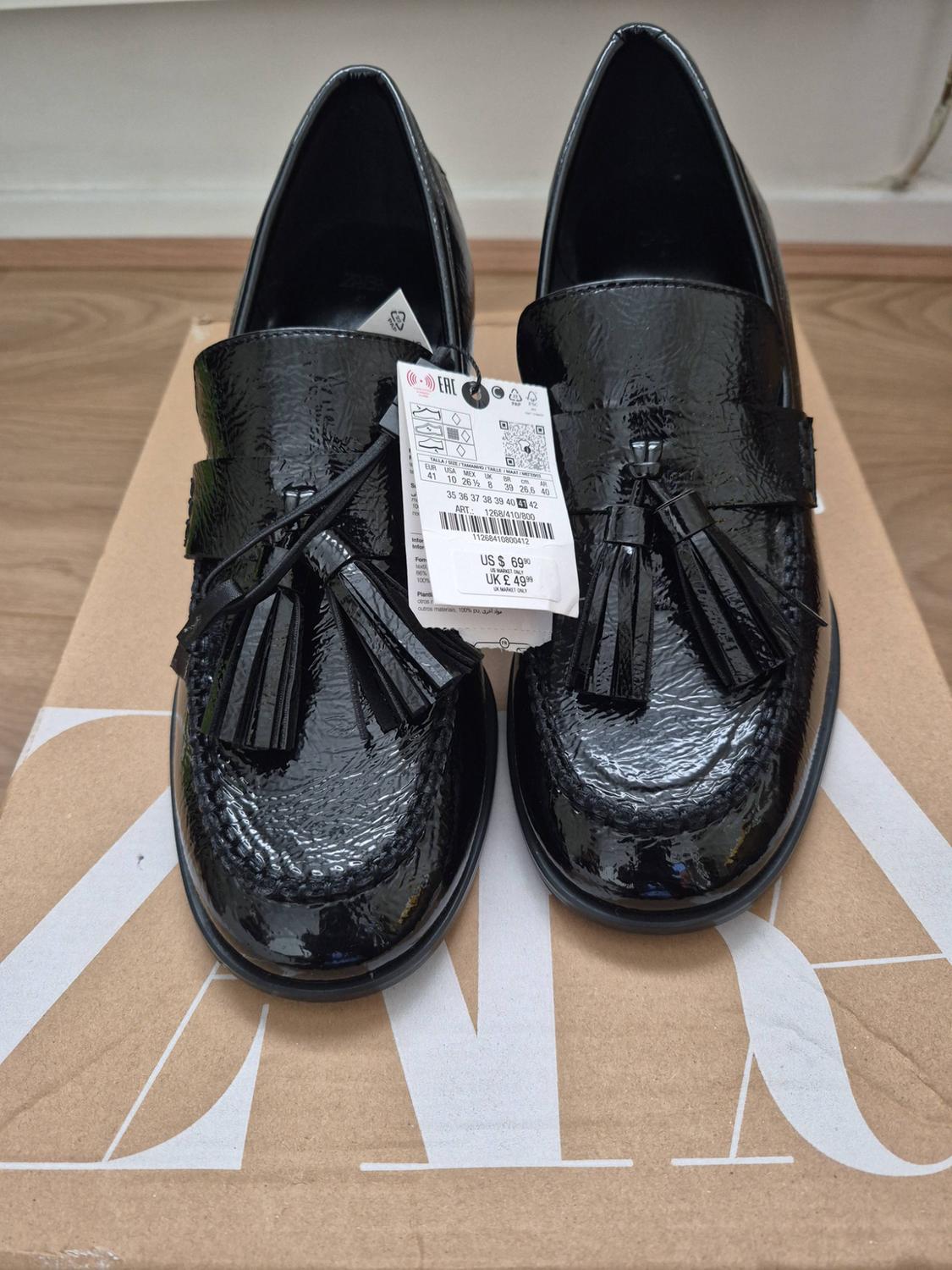 Shiny black tassel loafers on a box with the text ZARA. Label with text: ART.: 1268/410/800, US $69.90, UK £49.99.