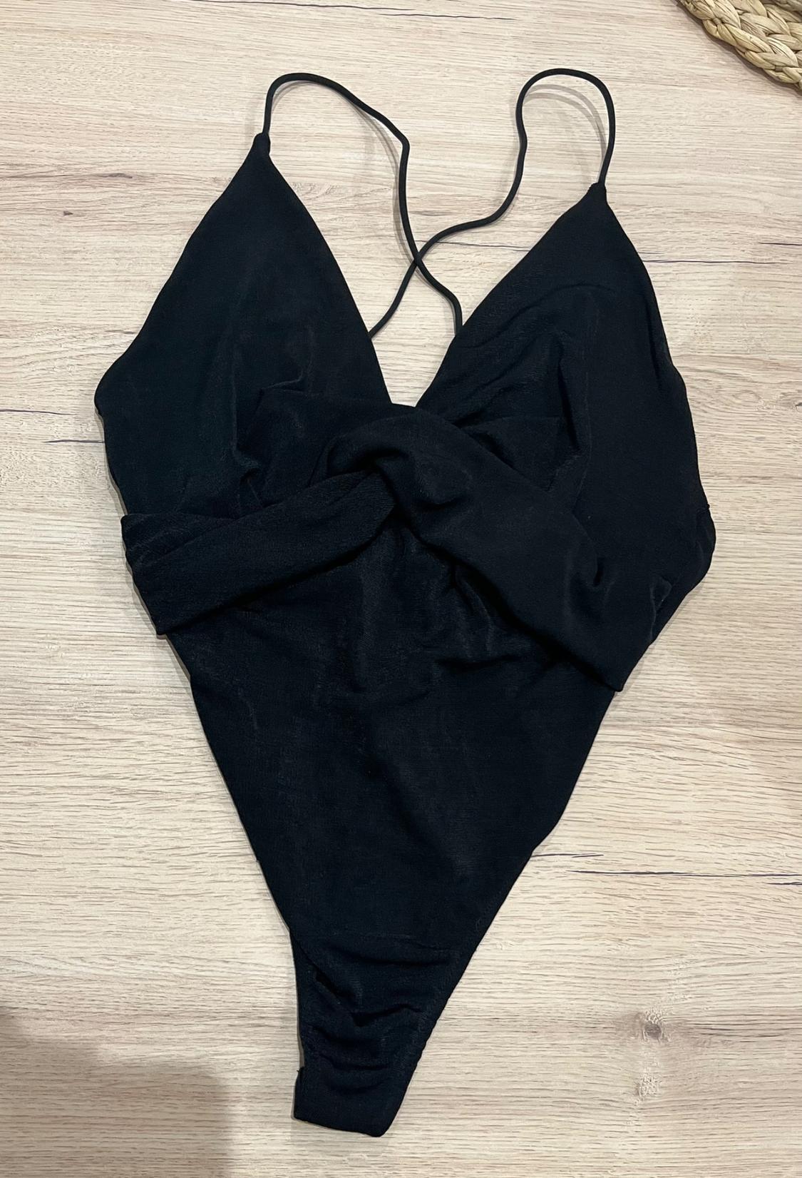 Black bodysuit with thin straps, a V-neckline and a knot detail at the front, presented on a light wood surface.