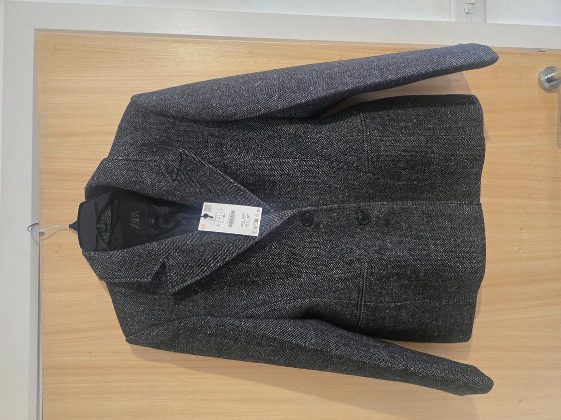Grey wool jacket hanging on a light wooden door, with a visible label that reads "Zara".