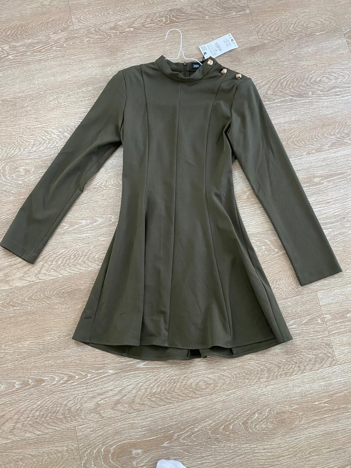 Olive green long-sleeved dress with gold buttons on the shoulder, hanging on a hanger over a light wood floor.
