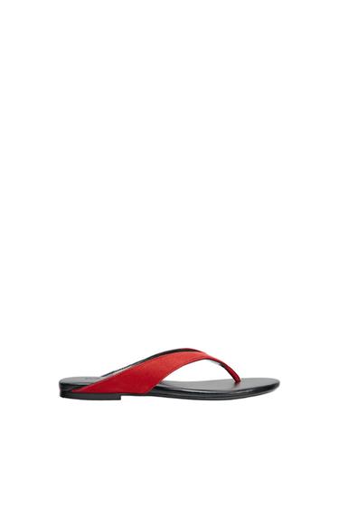 Minimalist composition of a flat sandal with a black sole and a red toe post, viewed in profile against a white background.
