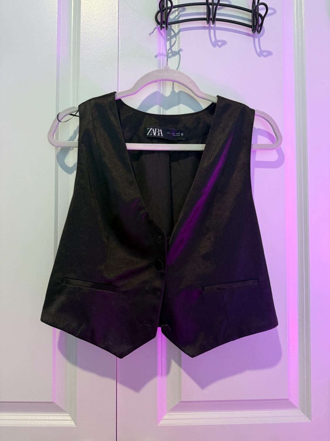 Black vest hanging on a white hanger in front of a door, illuminated with purple light. Visible label: "ZARA".