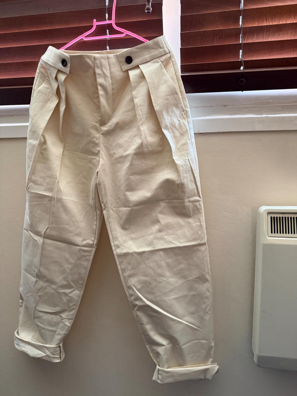 Beige tailored trousers with dark buttons and turn-up hems, hanging on a pink hanger in front of a window. Casual and relaxed style.