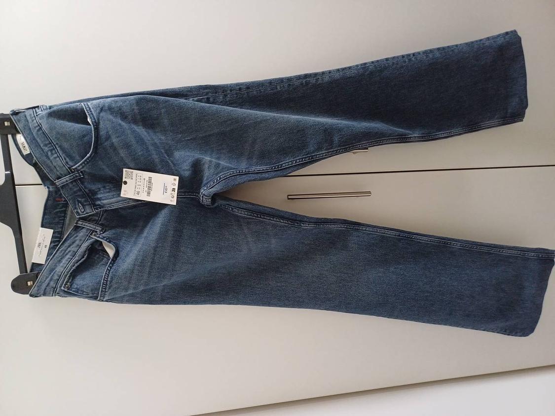 Blue jeans hanging on a black hanger, with visible tags on the waistband and leg, against a white background.