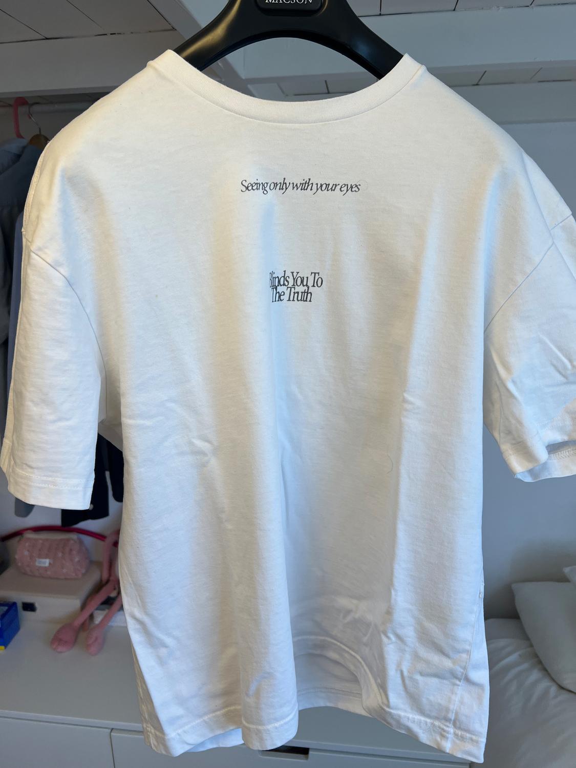 White t-shirt hanging on a black hanger. Text on the back: "Seeing only with your eyes blinds you to the truth". Room background.