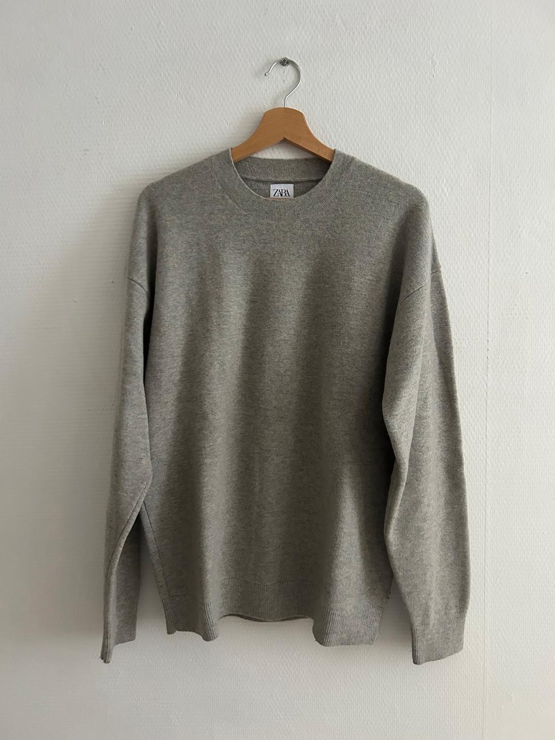 Grey long-sleeved jumper hanging on a wooden hanger against a white wall. Visible label with the text "ZARA".