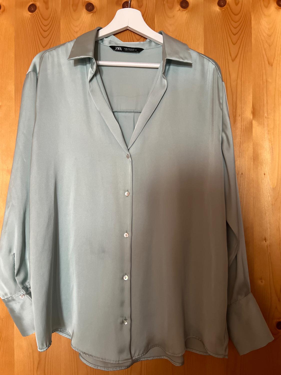 Light blue satin long-sleeved button-up shirt, hanging on a white hanger against a warm wooden background. Label with the text ZARA.