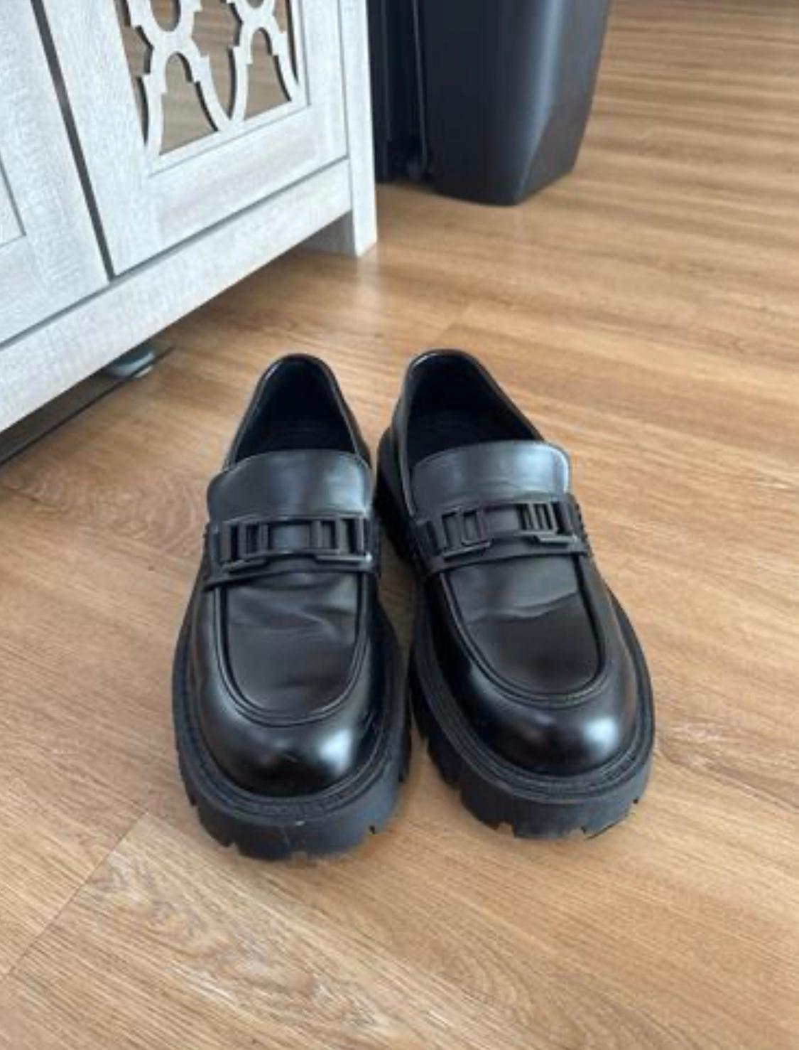 Black leather moccasins with a thick, ridged sole and a metallic detail on the upper, placed on a light wood floor.