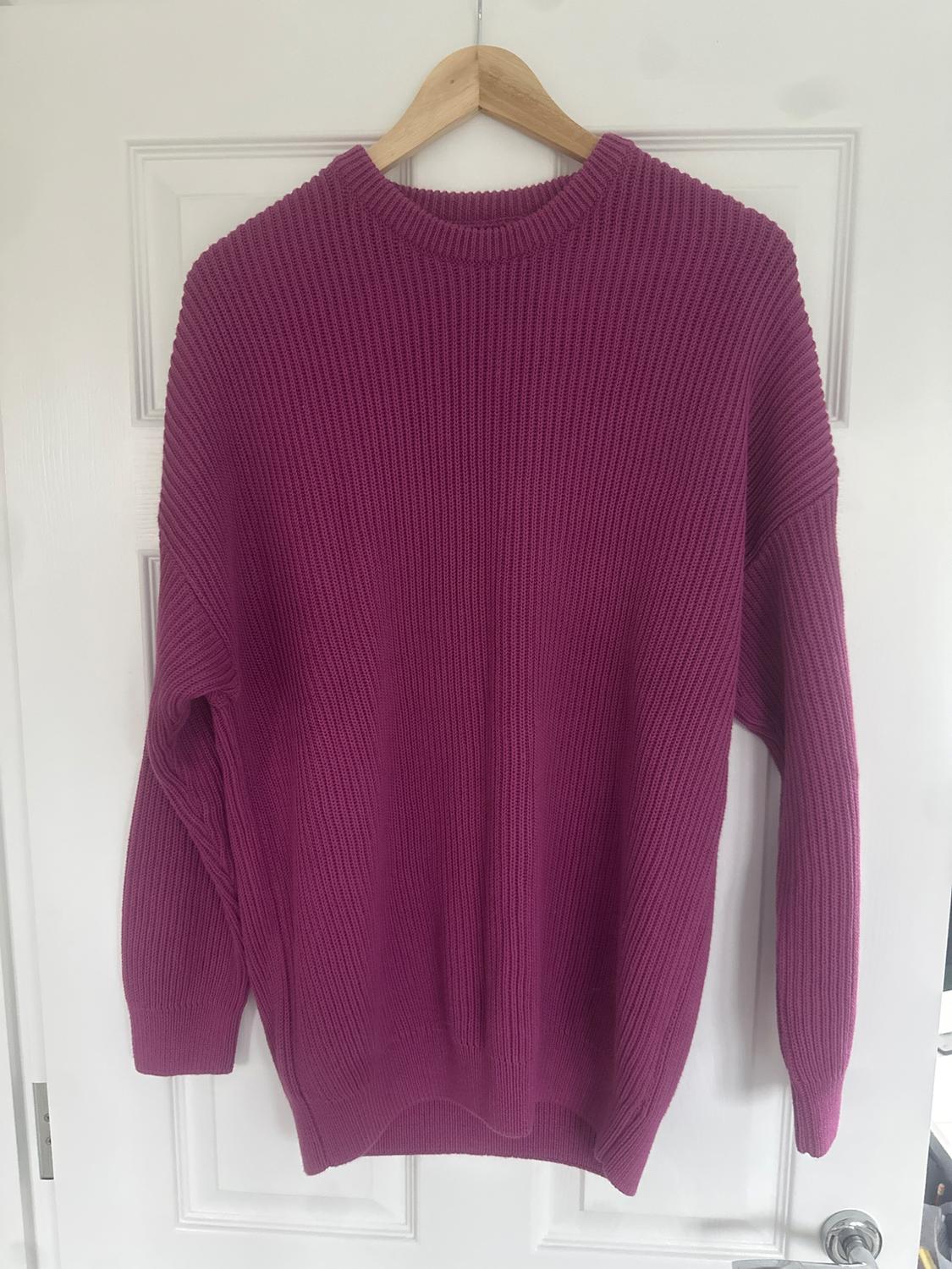 Fuchsia chunky knit jumper hung on a wooden hanger over a white door.