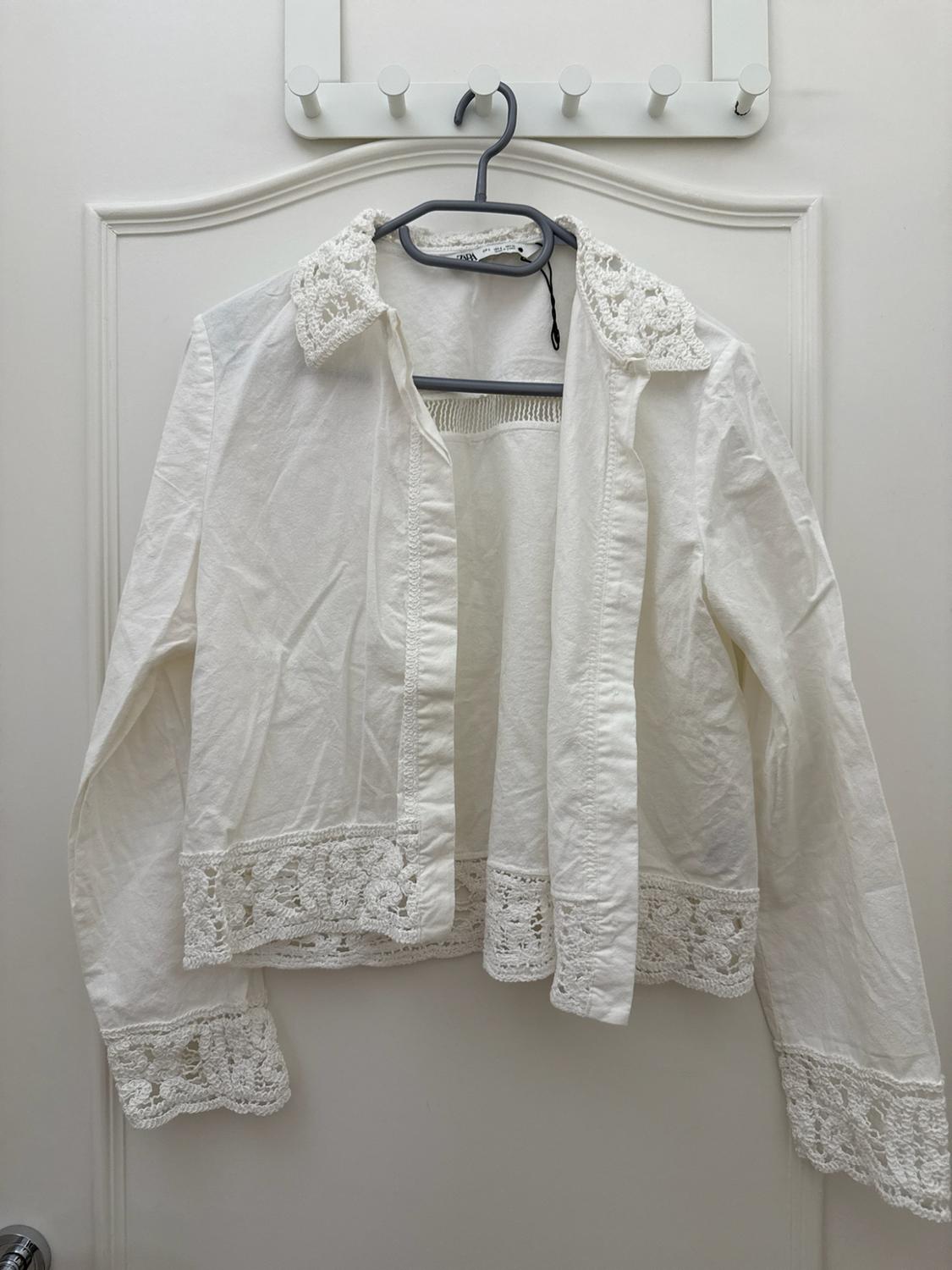Open white shirt with lace detailing on the collar, cuffs and hem, hanging on a hanger over a white door.