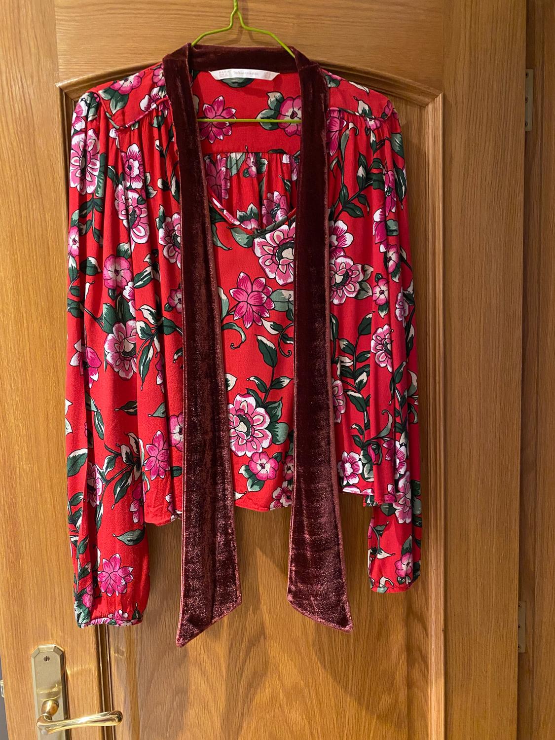 Red blouse with a pink and green floral print, hanging on a wooden door. Burgundy velvet bow detail.
