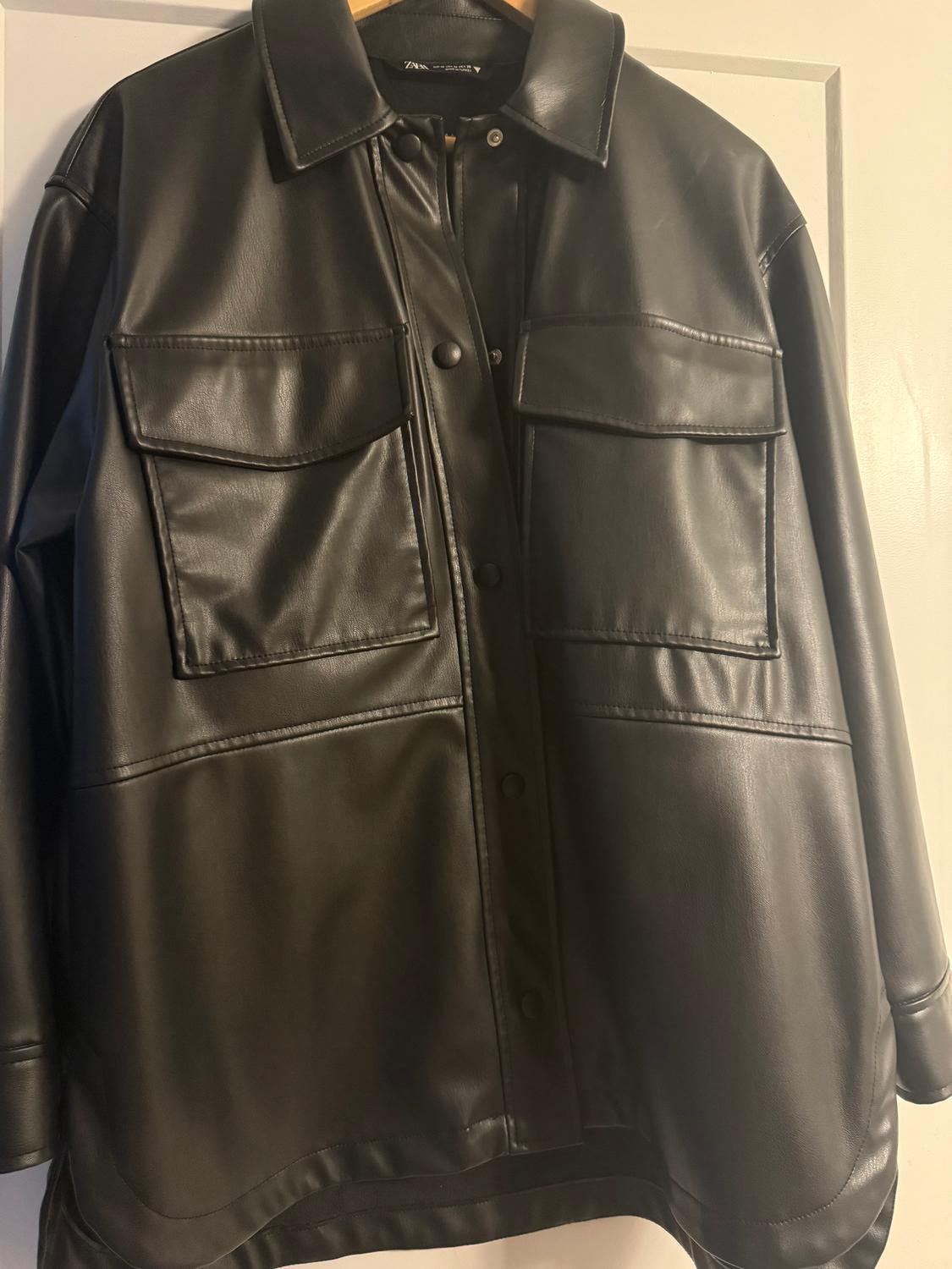 Black faux leather jacket with large pockets and front buttons, hanging on a hanger over a white door.