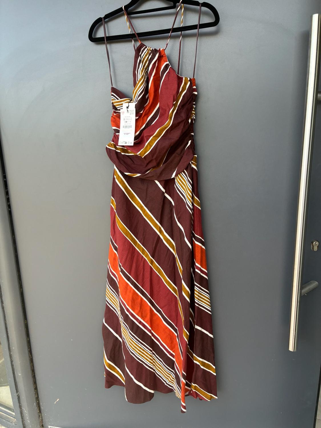 Long dress with diagonal maroon, orange, mustard and white stripes, draped detail and thin straps, hanging on a grey door.