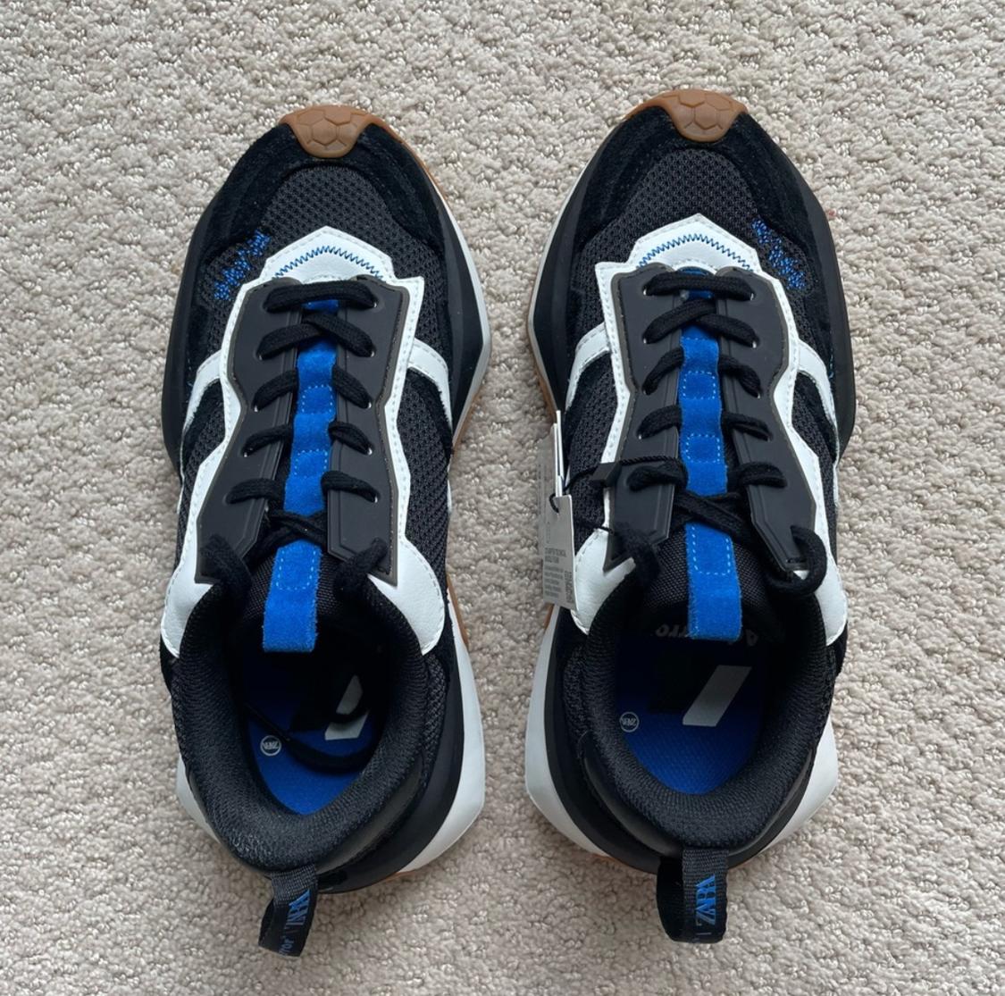 Black athletic shoes with white and blue accents, seen from above on a beige carpet. Black laces and a brown sole.