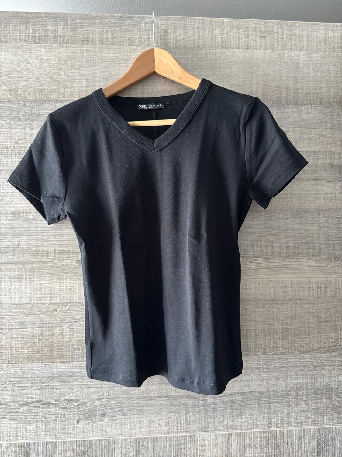 Black short-sleeved V-neck T-shirt hanging on a wooden hanger against a grey wooden background. Label with the text ZARA.