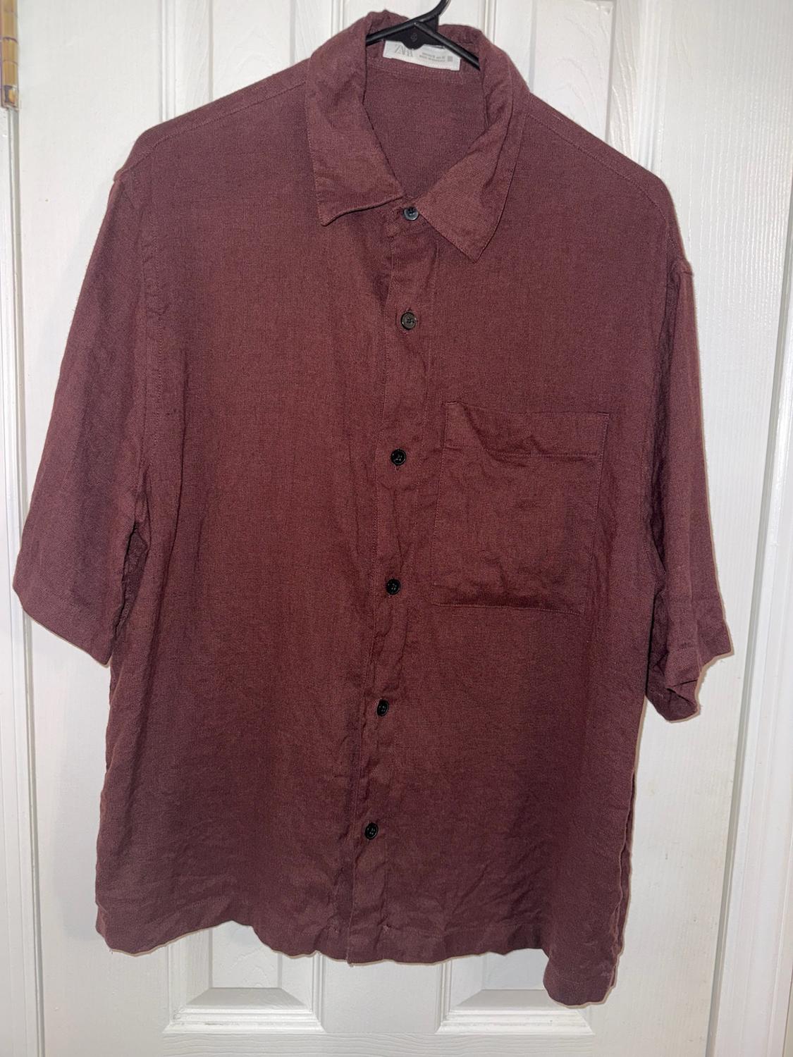 Short-sleeved maroon shirt with buttons and a chest pocket, hanging on a black hanger in front of a white door.