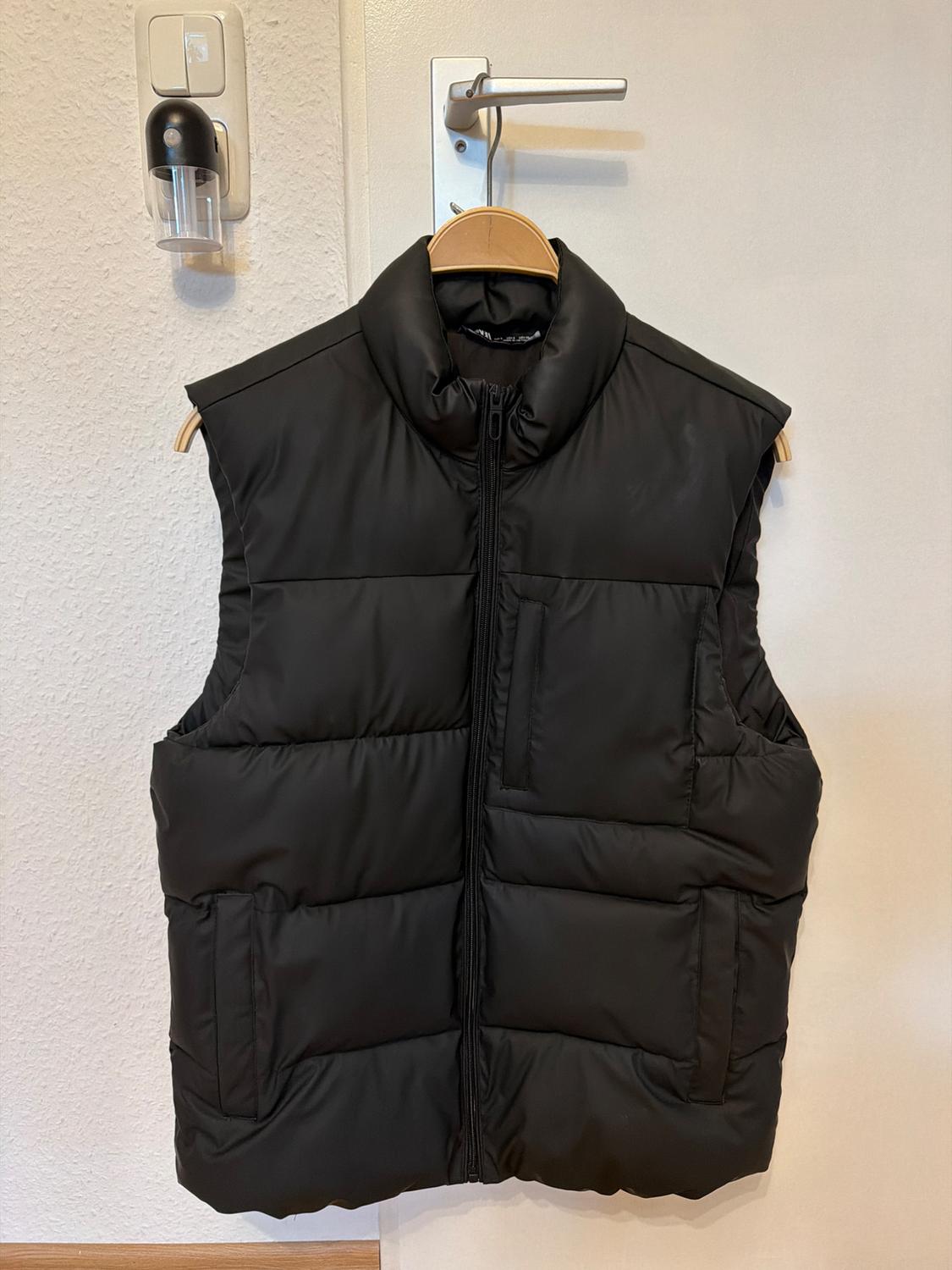 Black padded gilet hanging on a wooden hanger over a white door, with a dispenser on the wall in the background.
