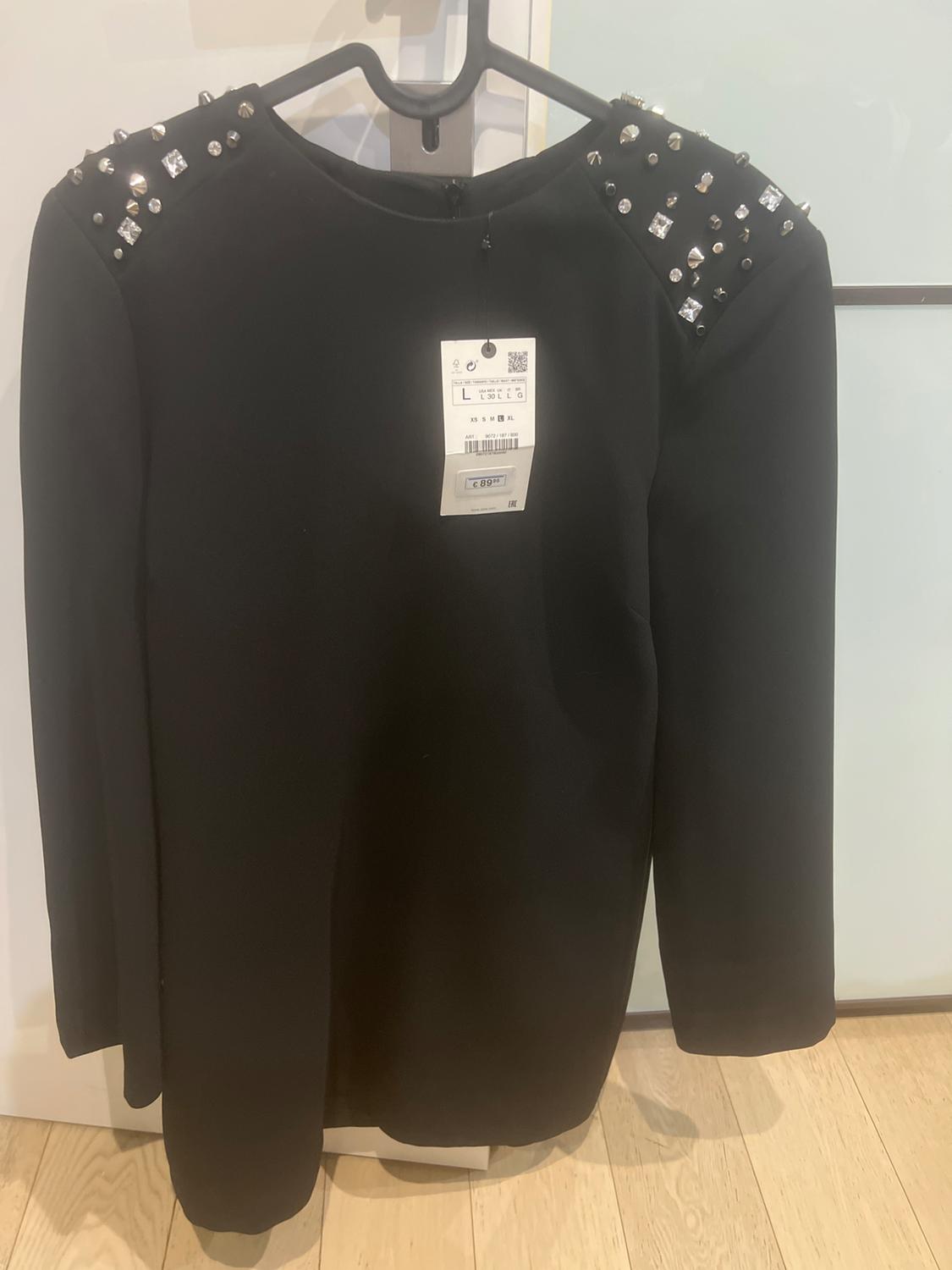 Black dress with long sleeves and shoulders adorned with studs and sparkling stones. Visible tag with barcode and price.