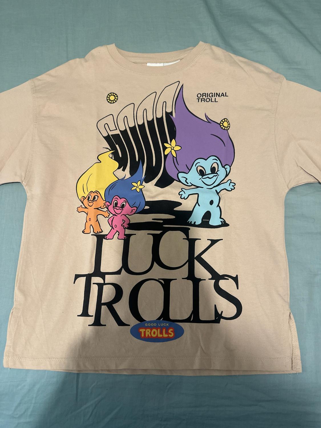 Beige short-sleeved T-shirt with a Trolls print and the text ORIGINAL TROLL, GOOD, LUCK TROLLS, GOOD LUCK TROLLS on blue fabric.