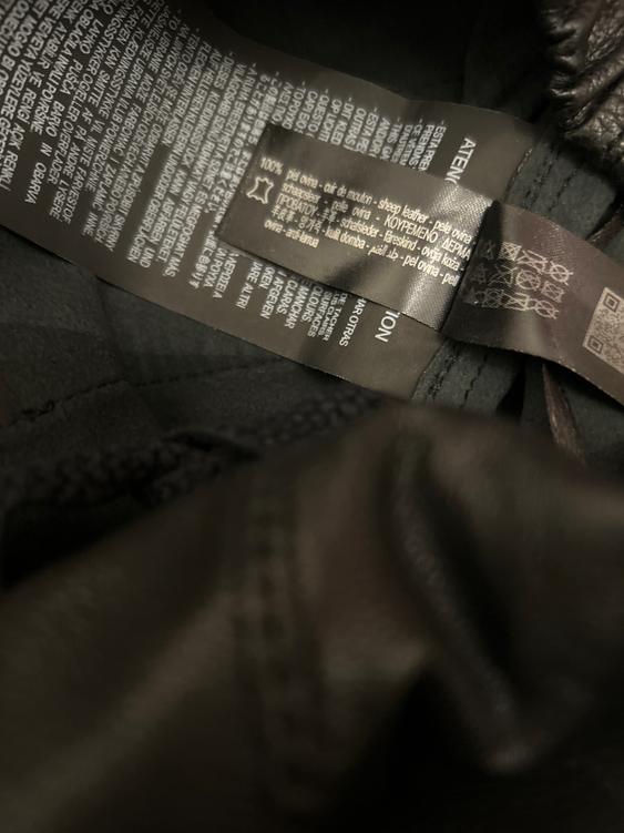 Close-up of clothing label with multi-language text, including "100% sheepskin". Dark background and visible fabric texture.