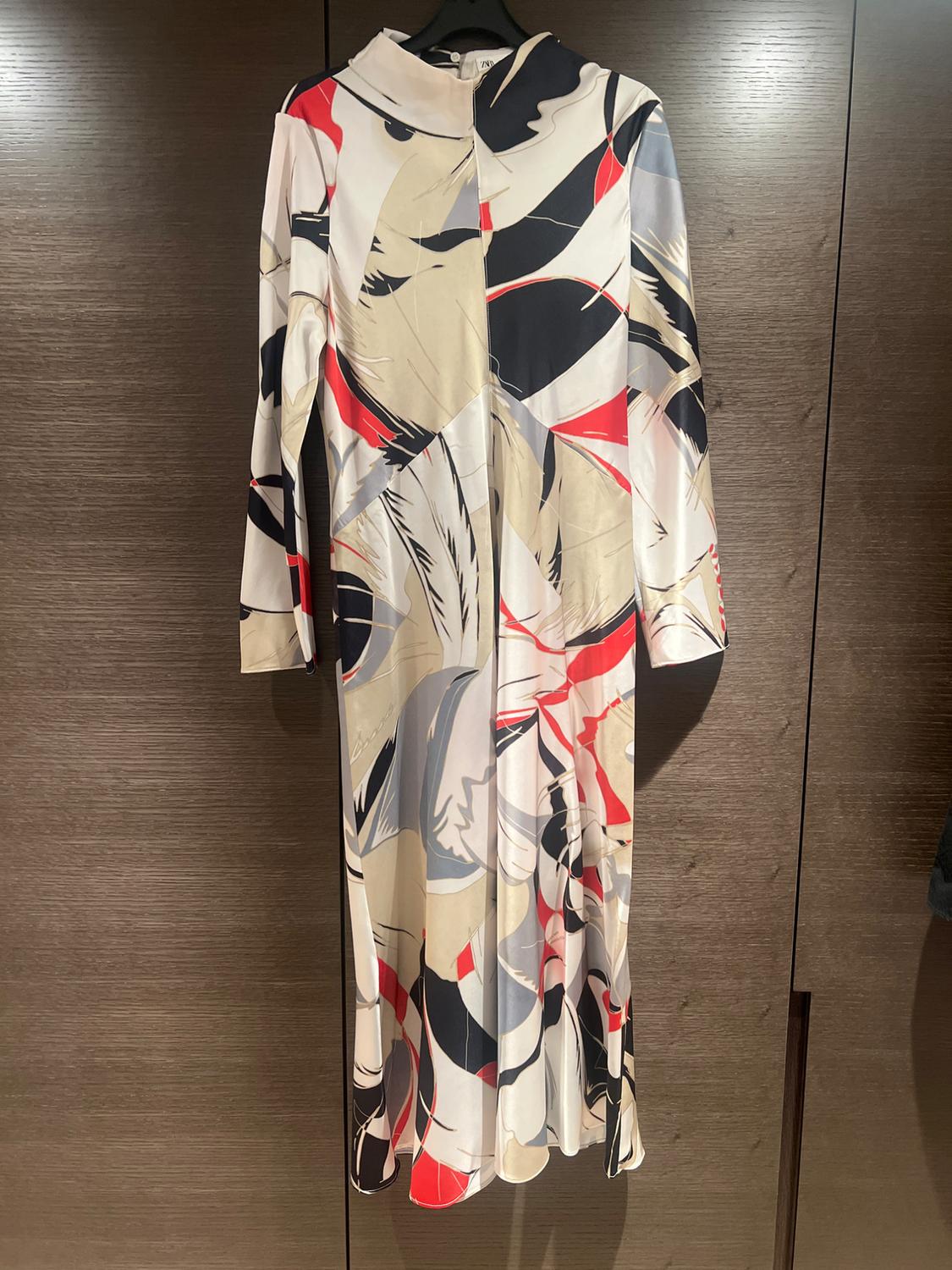 Long dress with an abstract print in beige, black, red, and white tones, hanging on a hanger against a dark wooden background.