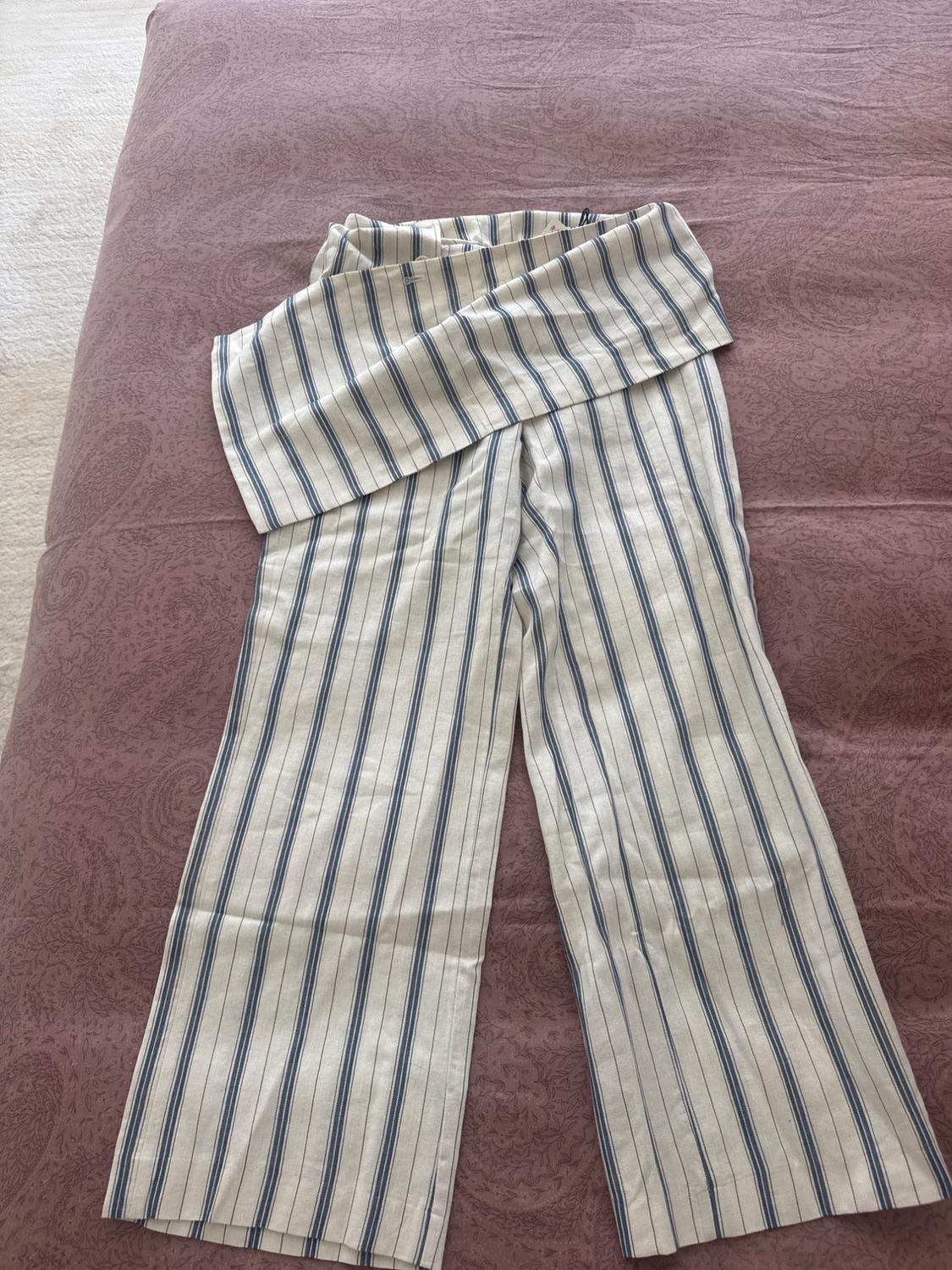Wide-leg trousers with blue and white vertical stripes and an asymmetrical folded waistband, laid out on a textured pink bedspread.