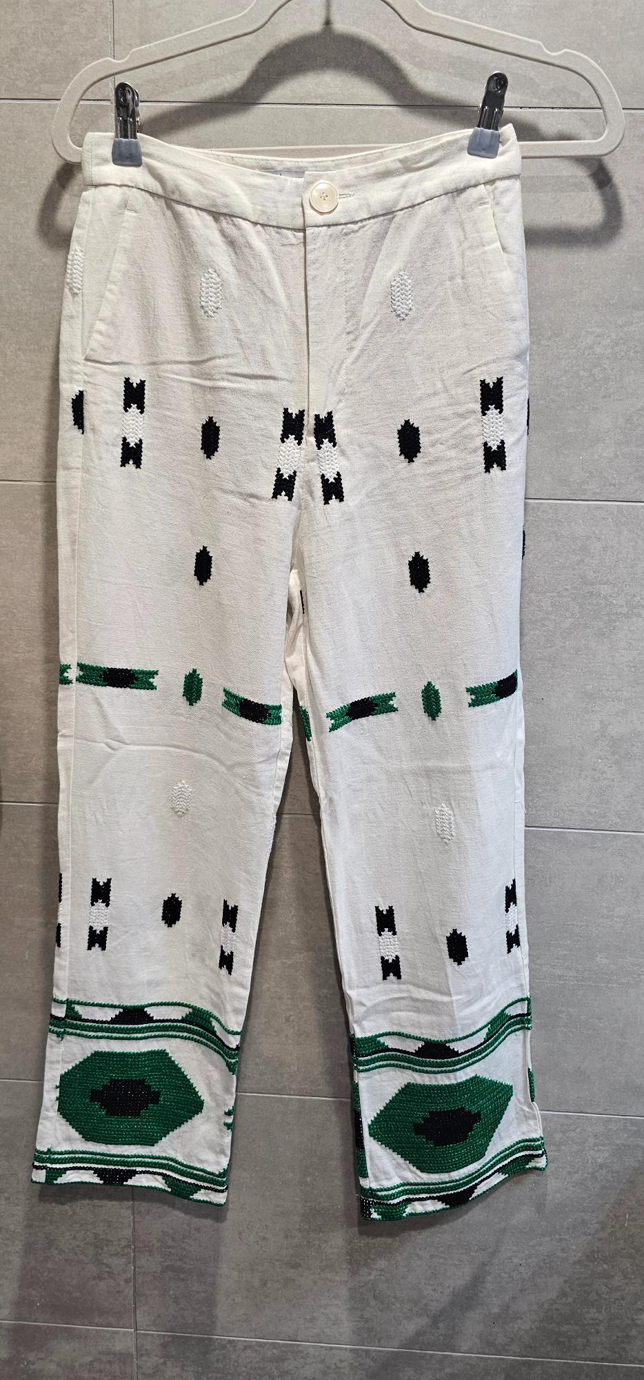 White trousers with geometric patterns in black and green, larger at the hem. Hanging on a hanger against a grey tiled wall.