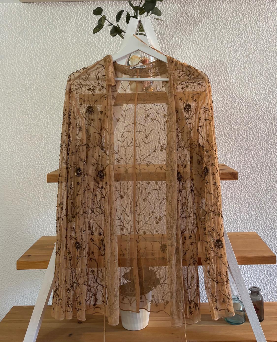 Sheer kimono embroidered in brown tones hung on a white hanger against a textured wall and wooden shelving background.