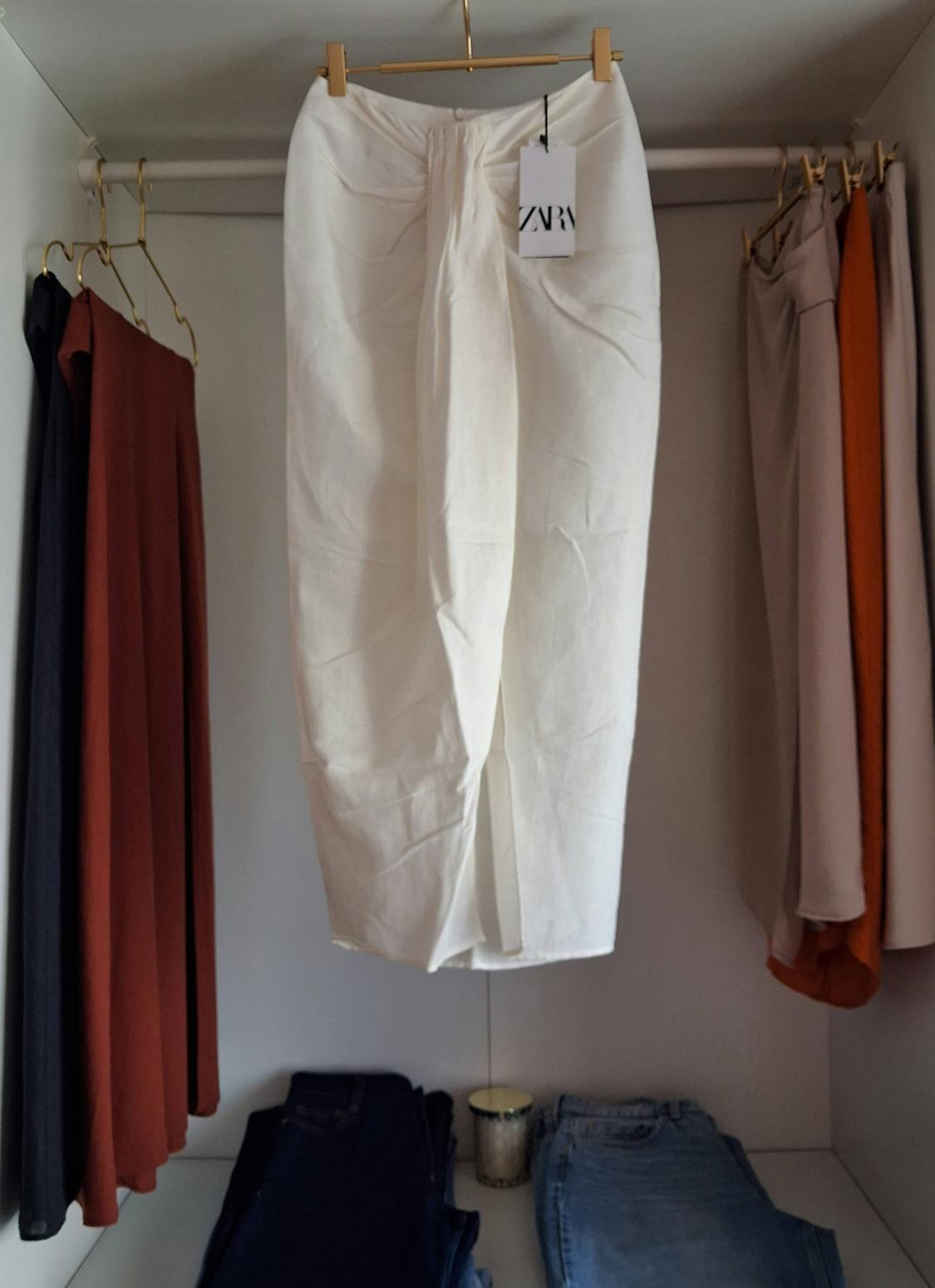 White skirt with a front knot hanging in a wardrobe. Label with the text ZARA. To the sides are hanging garments and folded jeans below.