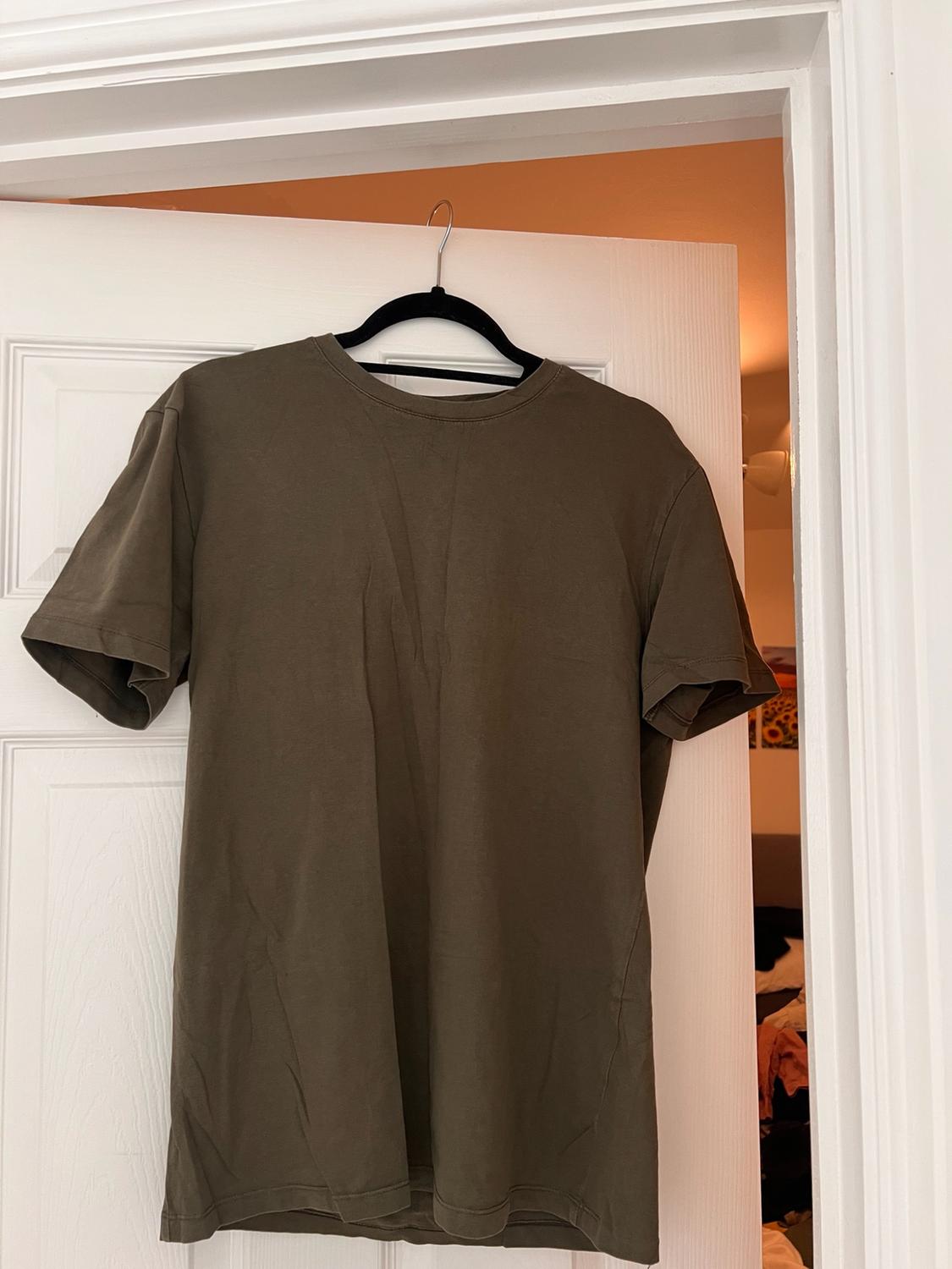 Olive green short-sleeved T-shirt hanging on a black hanger over an ajar white door, with a bedroom in the background.
