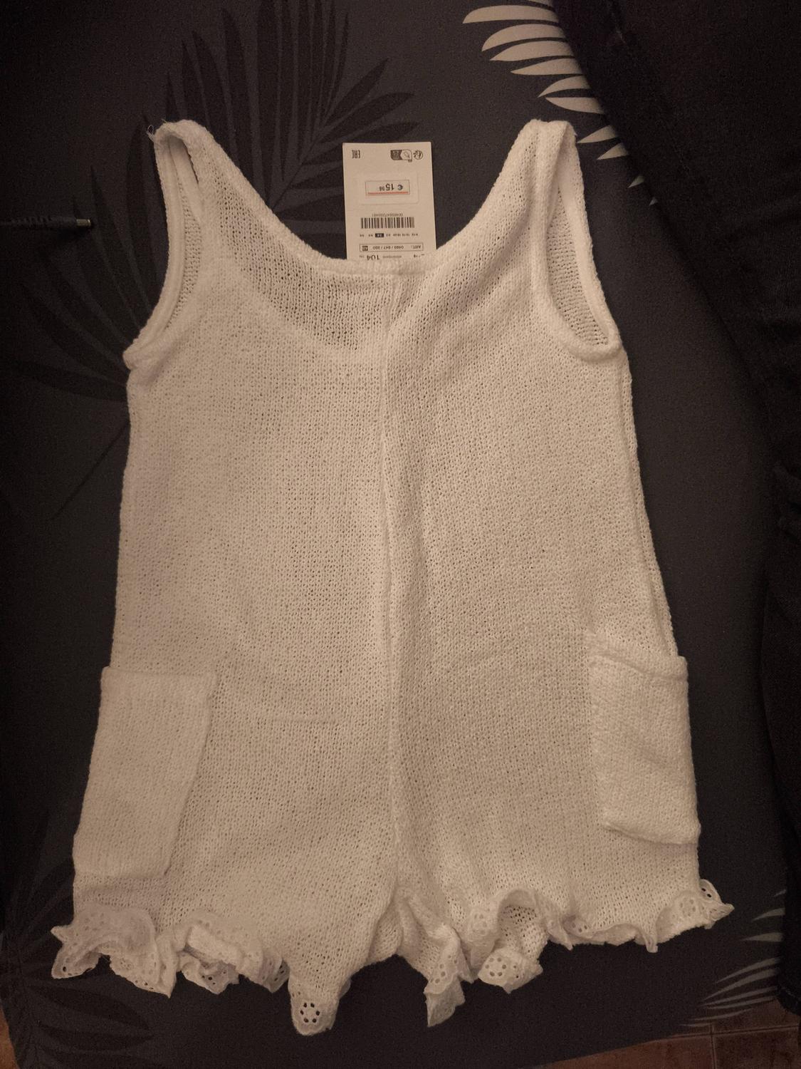 White knitted jumpsuit with straps and front pockets, on a dark surface with a leaf design. Visible label on the collar.