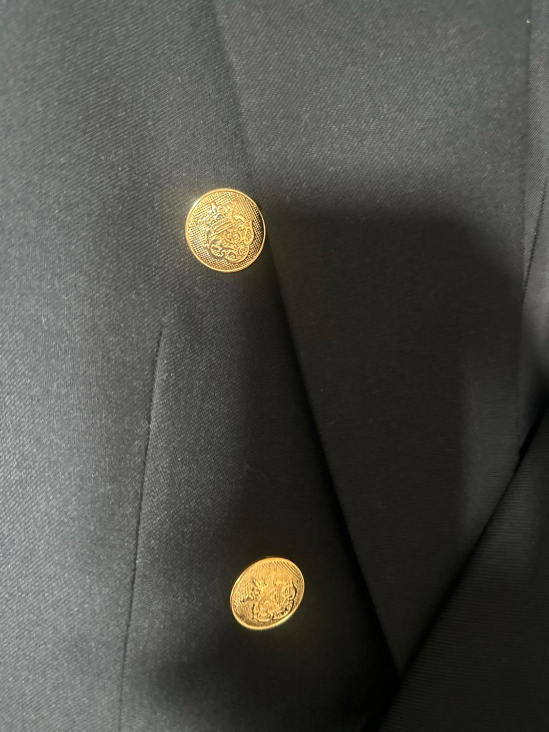 Close-up of a dark jacket showing two gold buttons with an embossed shield design, stitching and the texture of the fabric.