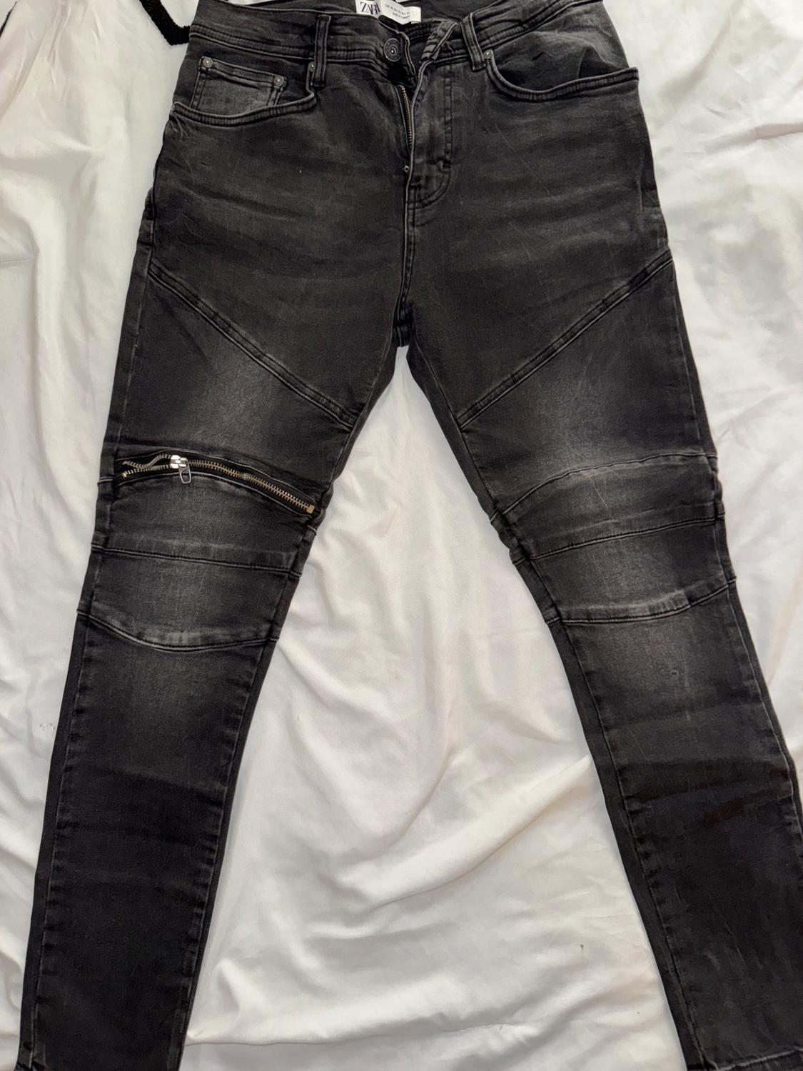 Black skinny pants with diagonal seam details and a zipper at the knee, on a wrinkled white background.