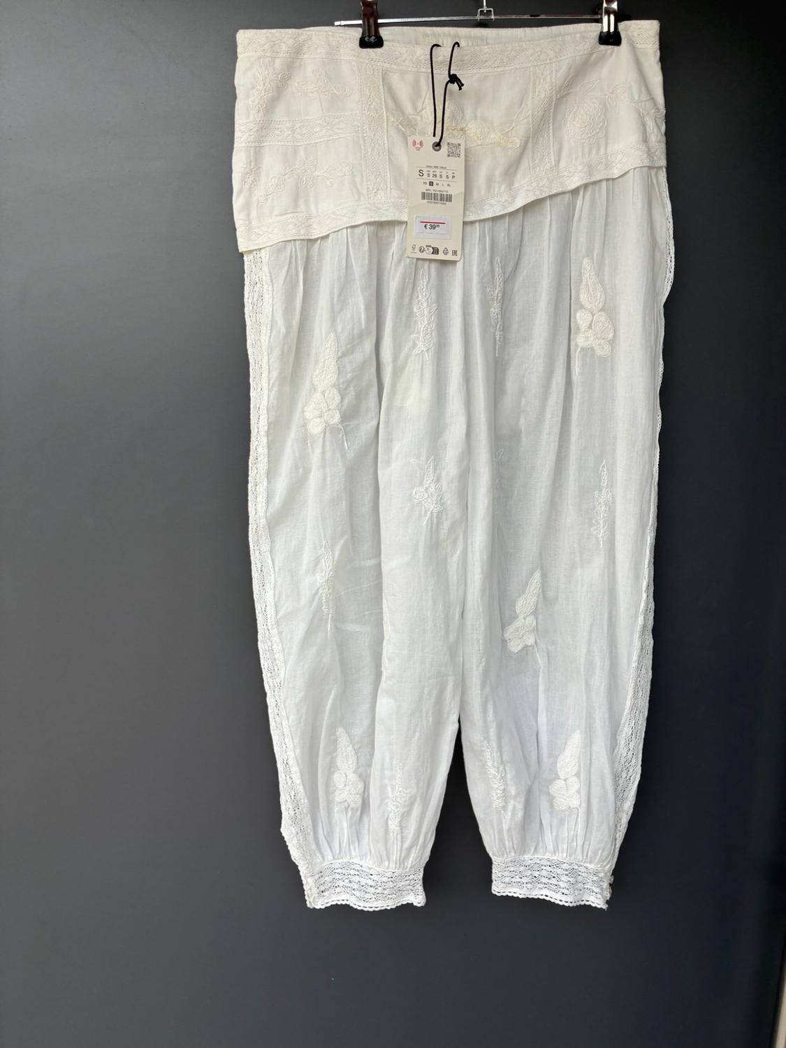 Wide white trousers hanging on a hanger against a grey background. They feature floral embroidery, lace details at the hems and a visible label.