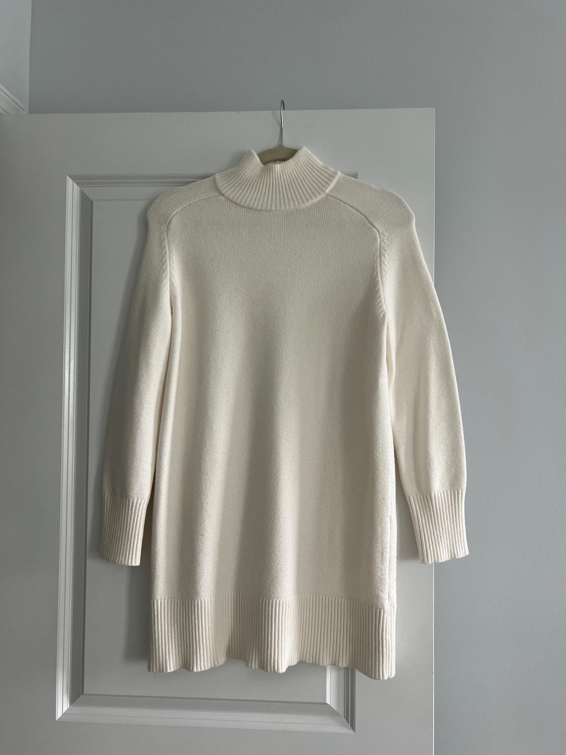White turtleneck sweater hanging on a white door. Simple design with long sleeves and ribbed detailing at cuffs and hem.