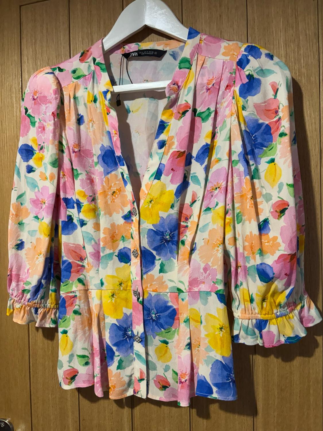White dress with a multicoloured floral print, buttons, and ruffled sleeves, hanging on a hanger against a wooden background. Text ZARA.