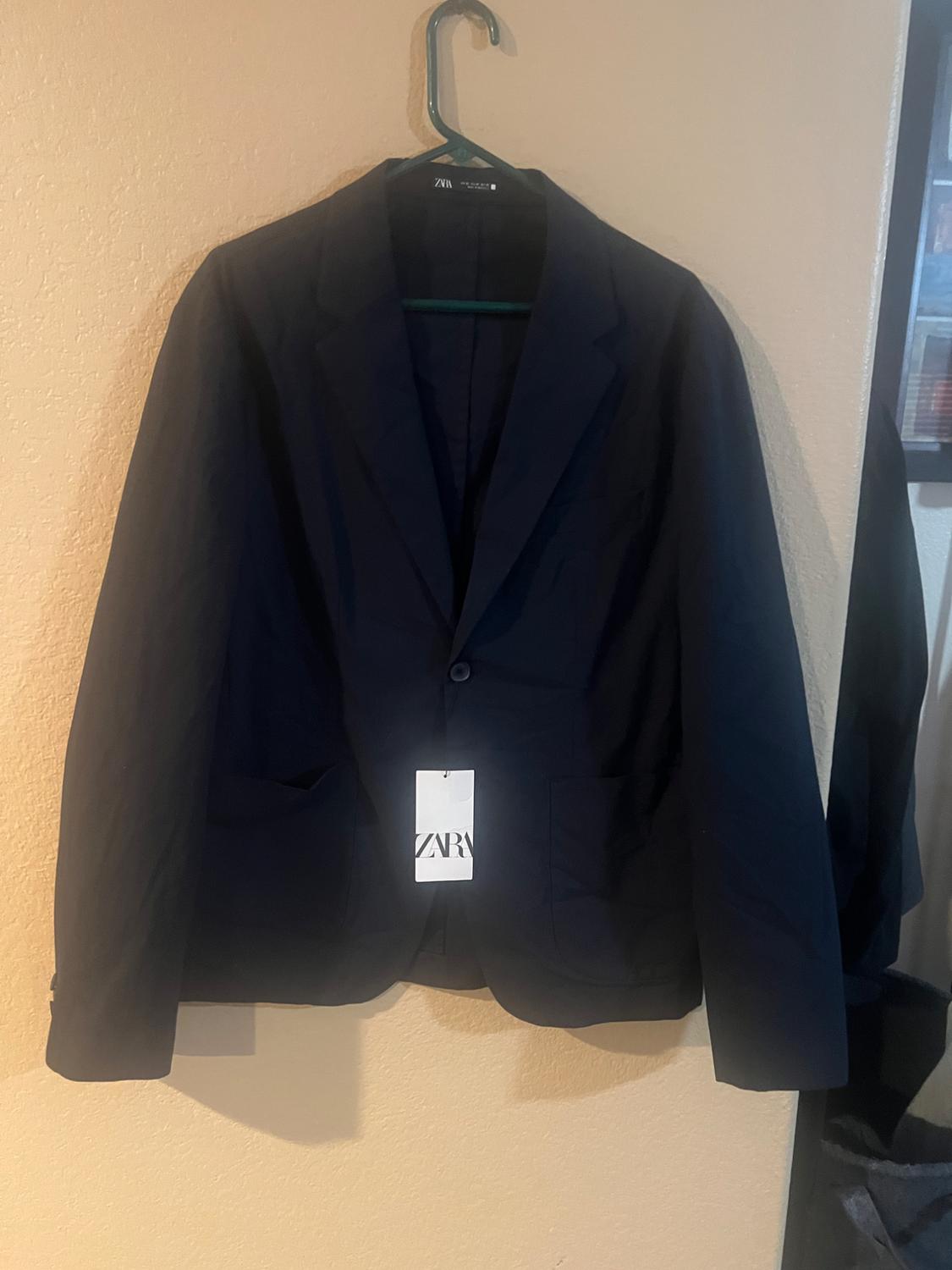 Navy blue jacket hanging on a green coat hanger, with a visible Zara label, beige background and part of a black garment on the right side.
