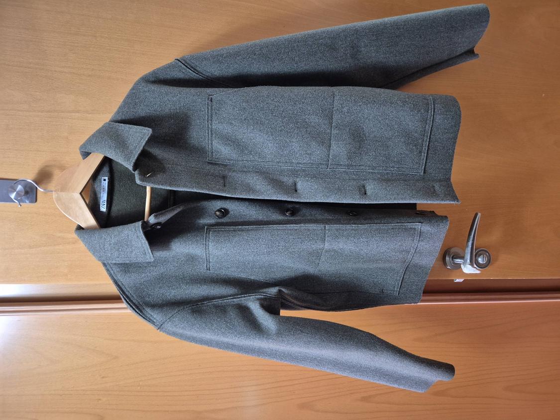 Grey wool jacket hanging on a wooden hanger over a light wooden door, with dark buttons and front pockets.